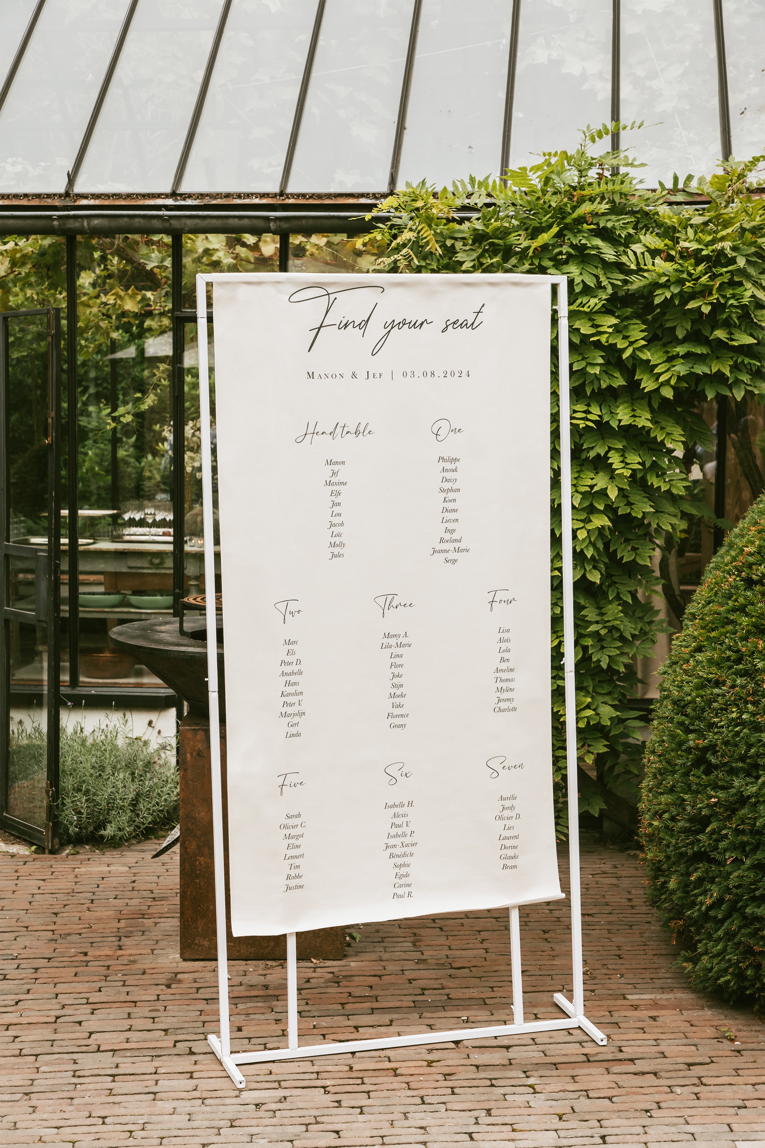 Plan de table en lin personnalisé avec le titre Find your seat, suspendu sur une structure blanche dans un joli décor verdoyant