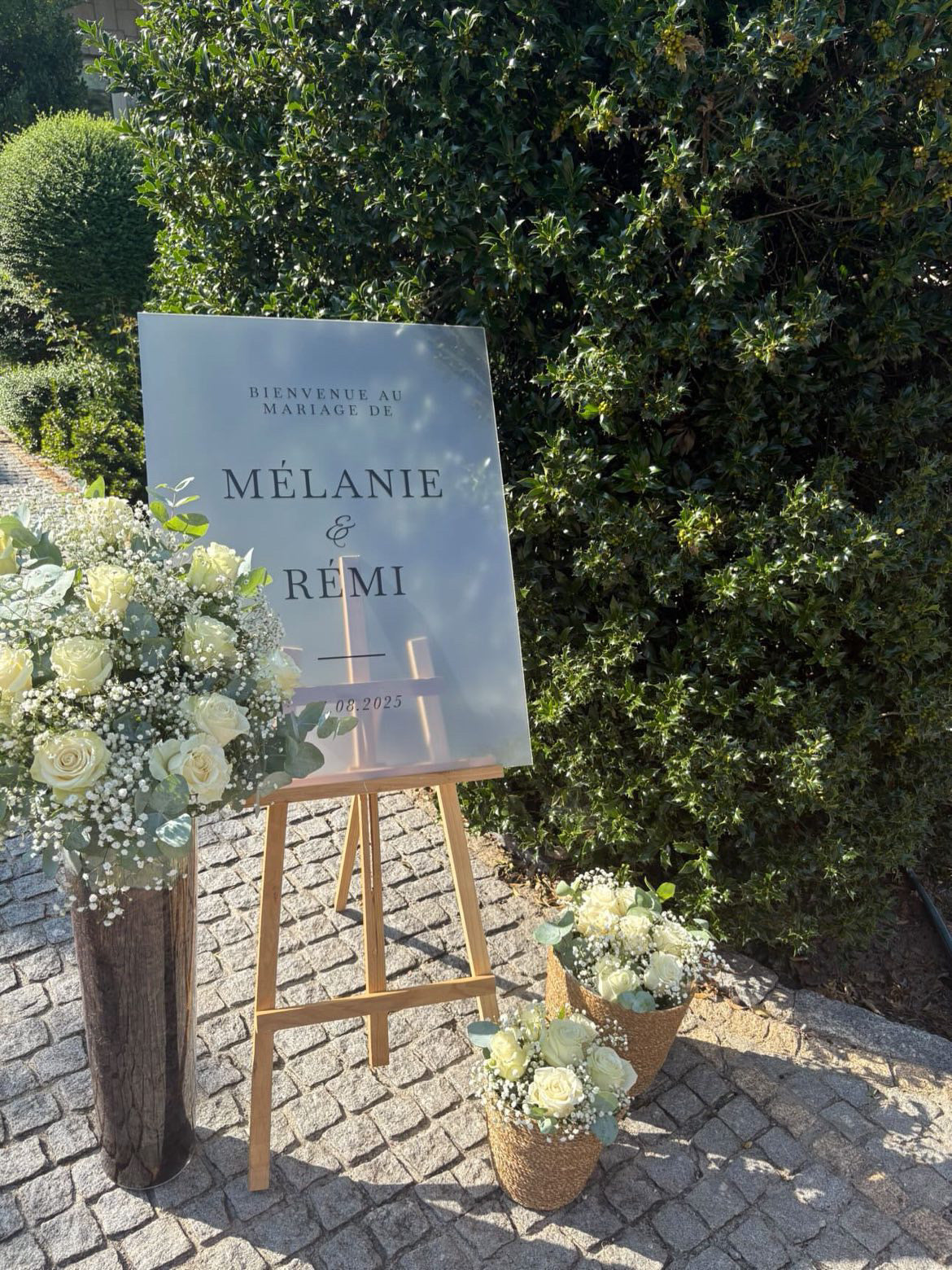 Panneau de bienvenue en plexiglass givré entouré de fleurs, installé en extérieur pour un mariage moderne.